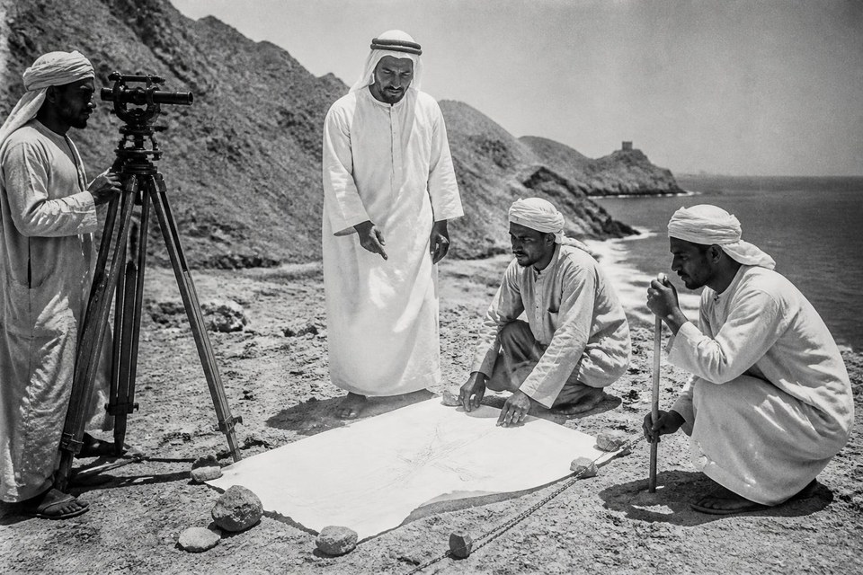 1952, East-coast road alignment, Fujairah, United Arab Emirates: Black-and-white photograph of Emirati surveyors reading a brass theodolite on the Fujairah east-coast road alignment in 1952, with the Hajar mountains and the Gulf of Oman behind them. — Avanti Holding, Foundations in Fujairah.