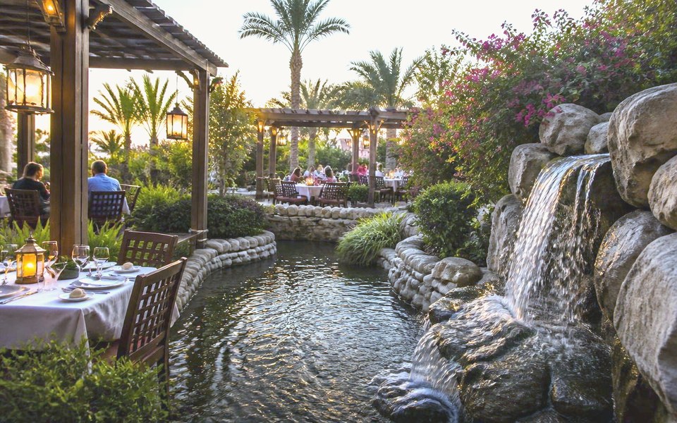 2027, Dubai, UAE: Visualisation of the planned JANNAH garden pavilion restaurant at dusk in Dubai, with diners on a shaded terrace beside cascading waterfalls and mature planting. — Avanti Holding, JANNAH.